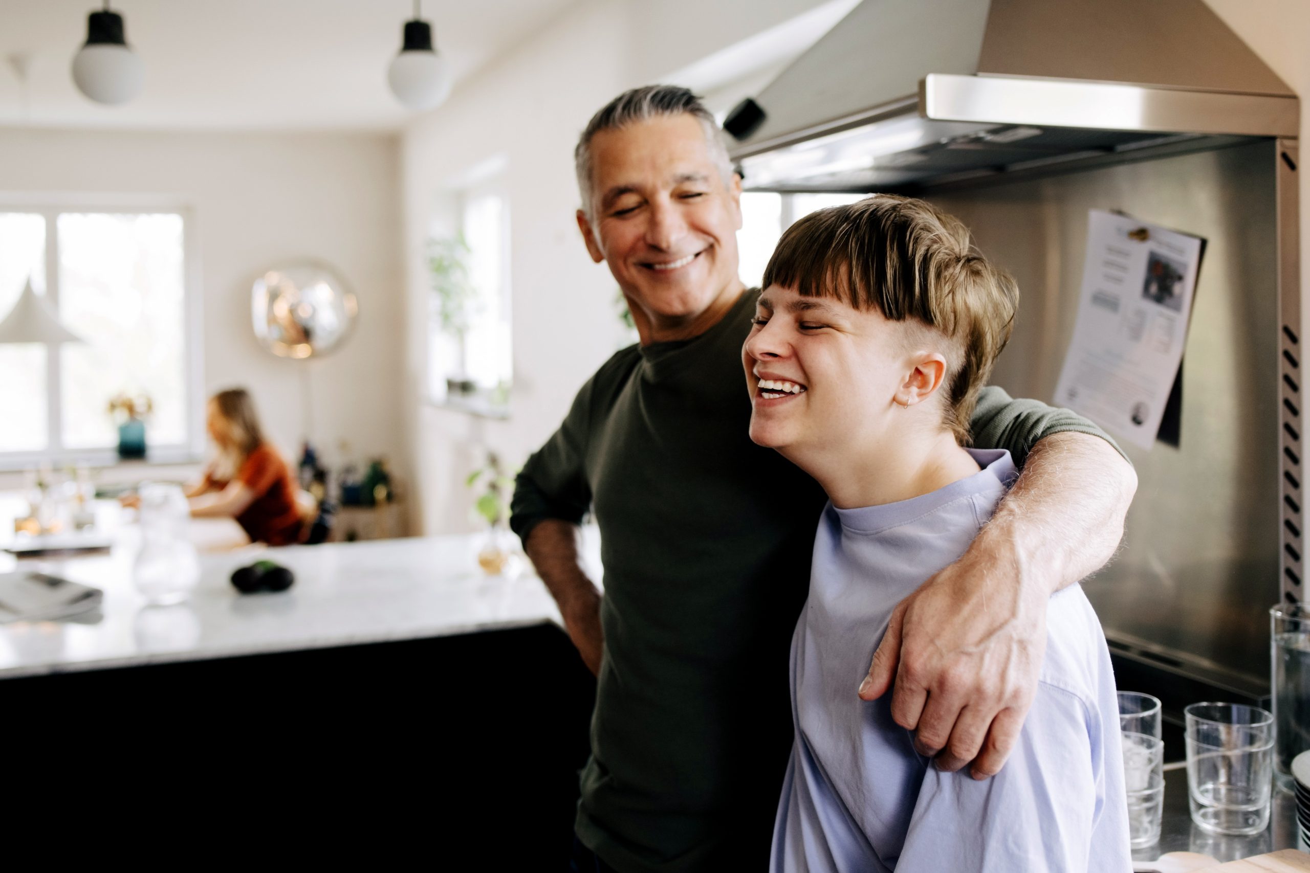 A joyful man and a young boy sharing smiles in a kitchen.